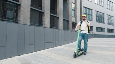 Sarı termal çantalı erkek kurye yiyecek teslimatı sokakta elektrikli scooterla gezerken akıllı telefon kullanıyor. Teslimatçı işçi çevrimiçi sipariş müşterisini teslim ediyor..
