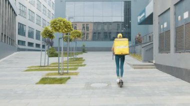 Yemek teslimatı yapan kadın kurye müşterilere sipariş ve paket götürmek için elektrikli scooter kullanıyor. Modern şehirde şık kentsel binalarla güneşli bir gün. Mal teslimatı ekolojik taşımacılık
