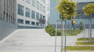 Sırt çantalı ve sarı şapkalı bayan teslimatçı şehir yolu boyunca elektrikli scooter kullanıyor. Kurye yemek teslimatı internetten elektrikli scooter siparişi ile sokaklarda geziyor.