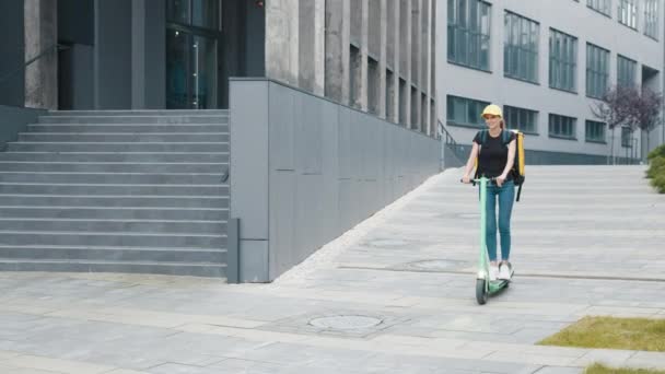 Pengiriman wanita dengan ransel naik skuter di jalan kota. Kurir wanita dengan ransel termal besar naik skuter listrik di jalan kota berpaling. Mengirim pesan online ke klien. — Stok Video