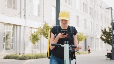 Teslimatçı kadın şehirde elektrikli scooterıyla dikilirken akıllı telefon kullanıyor. Şehre teslimatçı kadın ve telefondaki navigasyonu kullanarak restorandan yemek teslimatının adresini arıyor..