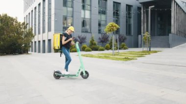 Sırtında büyük bir termal sırt çantasıyla kadın kurye portresi akıllı telefonda müşterisiyle şehir sokağında elektrikli scooterla konuşuyor. Teslimatçı kadın müşteriye sipariş getiriyor.