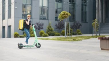 Kadın kurye yemek teslimatı, termal sırt çantalı cep telefonuyla sokakta elektrikli scooterla konuştuktan sonra müşteriyi arıyor. Teslimatçı kadın müşteriye sipariş getiriyor.
