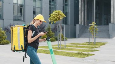 Sırt çantalı gülümseyen kadın akıllı telefona bakıyor. Şapkalı bayan teslimatçı akıllı telefondan sırayla bakar ve mutlu bir gülümsemeyle elektrikli scooter kullanır. Mal teslimatı ekolojik taşımacılık