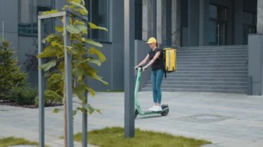 Gülümseyen kadın kurye, sokakta elektrikli scooterla gezen sarı termal çantalı yiyecekleri getiriyor. Pozitif dağıtım işçisi, çevrimiçi müşteri siparişi teslimatı.