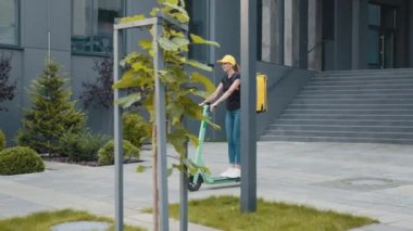 Genç kurye e-scooterına biniyor. Kadın paket teslimatçısı sokakta sarı sırt çantasıyla boş bir kaldırımda elektrikli scooter kullanıyor. Mal teslimatı.