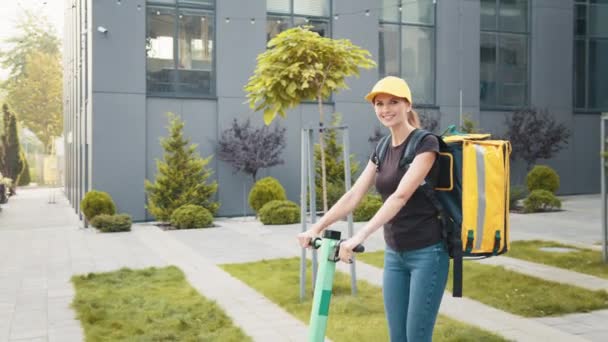 Uma jovem garota de entrega com um saco amarelo em suas costas monta uma scooter elétrica na rua no início da manhã. Trabalha em um serviço de entrega, com uma mochila amarela e um boné de beisebol amarelo — Vídeo de Stock