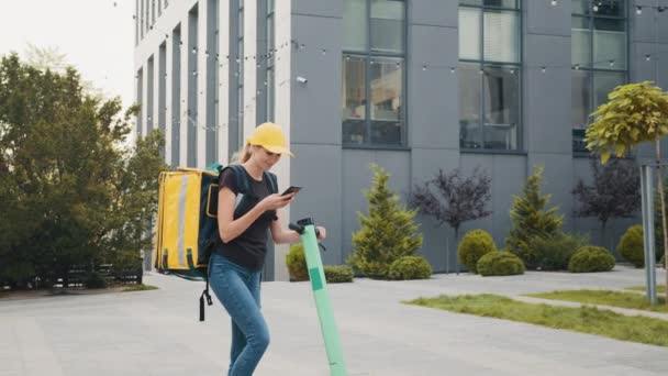 Zustellerin mit Smartphone in der Stadt mit Elektro-Schieberoller. Essenslieferung Kurier mit Thermobeutel und Roller mit Navigation im Telefon auf der Suche nach Adresse der Essenslieferung — Stockvideo