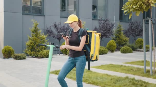 Deliveryman empleado trabajador entregar cliente de pedidos en línea. Entrega de comida de mensajero femenino con mochila térmica amarilla mira alrededor de la calle con scooter eléctrico utiliza navegador de teléfonos inteligentes — Vídeos de Stock