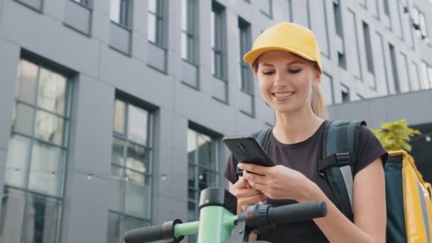 Entrega de comida mensajero mujer con bolsa térmica y scooter eléctrico. Entrega mujer con mochila térmica sosteniendo teléfono móvil esperando siguiente orden de pie con scooter eléctrico en la calle urbana — Vídeo de stock