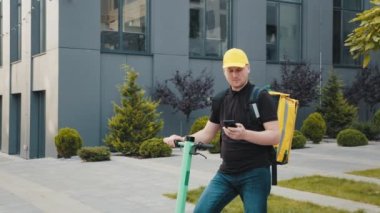 Erkek kurye bir restorandan yemek teslimatı için cep telefonu arıyor. Yemek teslimatçısı şehirde elektrikli scooterla geziyor. Çevre dostu ulaşım