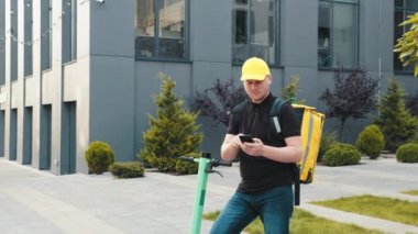 Teslimatçı işçi müşteriyi online siparişle teslim ediyor. Sarı termal çantalı erkek kurye yiyecek teslimatı sokakta elektrikli scooterla geziyor. Akıllı telefon navigasyonu kullanıyor..