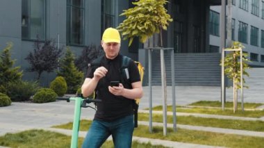 Bir kurye kameraya doğru işaret eden bir akıllı telefondan sipariş alır. Sırt çantası ve scooterlı teslimatçı telefondaki müşteri adresini arıyor..
