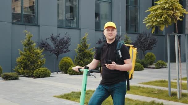 Jeune livreur avec scooter électrique utilisant un smartphone pour trouver le lieu de livraison. livreur avec sac à dos et un scooter est à la recherche de l'adresse des clients dans le téléphone. Il regarde la caméra. — Video