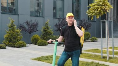 Sarı sırt çantalı yemek dağıtım elemanı siparişleri getirdi. Elektrikli scooterlı izotermal yiyecek kutusu olan erkek kurye evin girişine gelir ve müşteri çağırır. Hızlı teslimat.