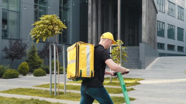 Usmívající se muž kurýr dodávky potravin s termálním batohem procházky ulici s elektrickým skútrem používá smartphone navigaci. Happy delivery man pracovník doručit on-line objednávky klienta — Stock video