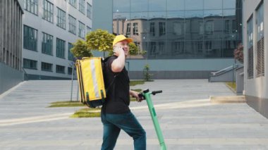 Sarı sırt çantası ve scooterlı teslimatçı telefondaki müşteri adresini arıyor. Kurye telefonundaki müşteri adresini kontrol ediyor, yemek teslimatı salgını. Hızlı teslimat.