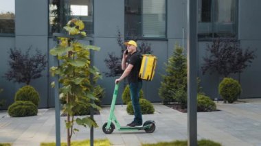Şehirde elektrikli scooterlı akıllı telefon kullanan teslimatçı. Termal sırt çantalı genç teslimatçı scooter 'ın yanında ve dışarıda akıllı telefon kullanıyor..