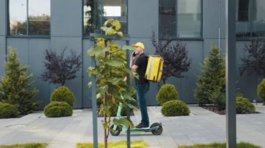 Termal sırt çantalı genç teslimatçı modern binanın yakınındaki scooter 'da akıllı telefon kullanıyor. Smartphone kullanan teslimatçı Scooter 'lı restoranlardan yiyecek getiriyor..