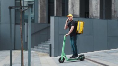 Elektrikli scooter ve termal sırt çantalı kurye yemek teslimatı akıllı telefon kullanarak müşteri siparişini onaylıyor. Elektrikli scooter kullanan bir adam telefonda konuşuyor.