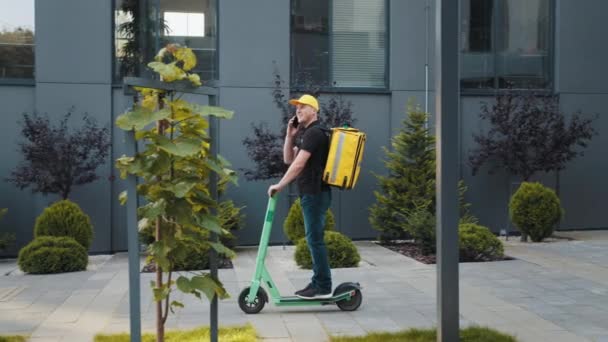 Şehirde elektrikli scooterlı akıllı telefon kullanan teslimatçı. Termal sırt çantalı genç teslimatçı scooter 'ın yanında ve dışarıda akıllı telefon kullanıyor.. — Stok video