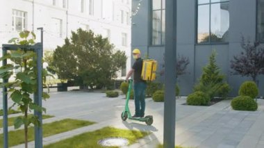Sarı sırt çantalı bir teslimatçı video çerçevesine elektronik motosikletle giriyor, etrafa bakınmayı kesiyor, adam sırtından vuruluyor. Karantina sırasında mükemmel atış..