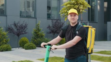Elektrikli scooterlı, sarı termal sırt çantalı ve kameraya bakan teslimatçının portresi. Müşteri evine ya da ofisine fast food getiren neşeli kurye. Ekspres Teslimat