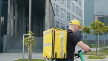 Beyaz erkek kurye E-scooter 'la müşteri evine ya da ofisine yiyecek getiriyor. Caddeye doğru giden çekici erkek teslimatçı. Restoran, kafe, süpermarket yemek teslimatı.