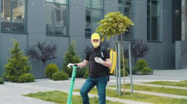 Şehir caddesinde yalıtımlı çantalı tıbbi maske takan teslimatçının orta boy fotoğrafını çekiyorum. Sırtında sarı bir sırt çantası olan teslimatçı telefonda bir müşteriyle konuşuyor..