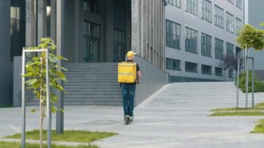 Sarı sırt çantalı bir teslimatçı şehirde yiyecek getiren elektrikli bir scooter kullanıyor. Şehir kurye servisi, kurye bisiklet sürüyor, video da arkadan çekiliyor..