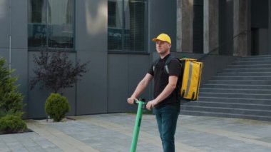 E-scooter 'a binip siparişi getiren yakışıklı beyaz bir teslimatçı. Çekici erkek kurye, internetten aldığı yiyecekleri getiriyor. E-ticaret, online alışveriş.
