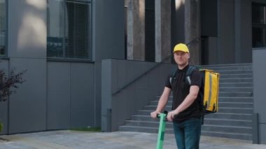 Sarı sırt çantalı bir teslimatçı şehirde yiyecek getiren elektrikli bir scooter kullanıyor. Erkek kurye, internet mağazasından aldığı yiyecekleri getiriyor. E-ticaret, online alışveriş.