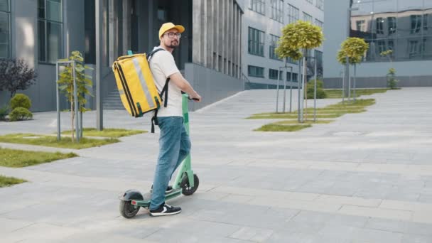 Portrait de l'homme livreur debout dans la rue et tournant face à la caméra. Coursier masculin avec sac à dos jaune et scooter électrique. Livrer le transport vert. Concept de livraison — Video