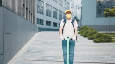 Sarı sırt çantalı ve koruyucu maskeli bir teslimatçı yemek teslimatı ve iş merkezi geçmişiyle şehirde elektrikli scooter kullanıyor. Karantina sırasında mükemmel bir atış. Konsept Teslimat.