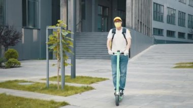Sarı sırt çantalı ve koruyucu maskeli bir teslimatçı yemek teslimatı ve iş merkezi geçmişiyle şehirde elektrikli scooter kullanıyor. Yiyecek kutusu olan kurye elektrikli scooterla geliyor.