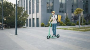 Teslimatçı koruyucu maske takıyor ve modern evlerin ya da büroların önüne mobiletli çanta, ekspres yemek teslimatı ve online alışveriş konseptiyle yiyecek paketi göndermeye hazır..