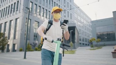 Satıcı akıllı telefon ekranına bakar ve caddede telefonla konuşur. Maskeli kurye, modern bir ofis binasının arka planındaki salgın sırasında yemek dağıtıyor..
