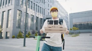 Maskeli kurye elinde kahve, pizza kutusu, güvenlik eldiveni takıyor ve içki ikram ediyor. Yakışıklı teslimatçı elinde bir yığın pizza kartonuyla. Pizza yiyen erkek kurye kameraya bakıyor..