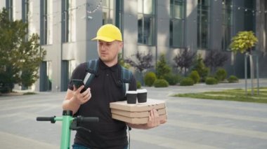 Portret Young kurye, pizza ve kahve teslimatçısı, termal sırt çantalı ve e-scooter 'lı. Eve hızlı ulaşım ekspresi. Çevrimiçi sipariş. Akıllı telefona bakan şapkalı teslimatçı.
