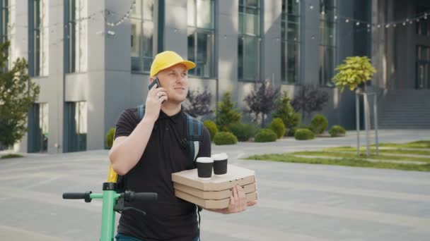 Portret młodego kuriera dzwoniącego przez telefon komórkowy do klienta na zewnątrz. Kurier dostarczający jedzenie i gorące napoje na zewnątrz. Facet używający smartfona na ulicy. Kurier pizzą i kawą — Wideo stockowe