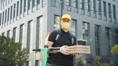 Eldivenli ve tıbbi maskeli yakışıklı beyaz bir teslimatçının portre fotoğrafı. Sokakta duruyor, kutuları tutuyor ve tutuyor. Erkek kurye pizza ve kahve veriyor. Modern bir ofise teslimat..