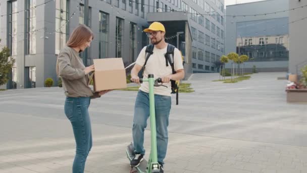 Correio entregando um pacote em uma scooter elétrica. Menina sorrindo abrindo uma caixa de papelão e puxa para fora sapatos. Entrega de uma loja de roupas online. Entrega de sapatos. Compras online. — Vídeo de Stock