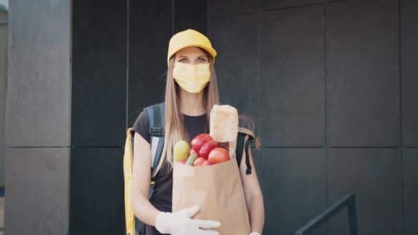 Porträt einer kaukasischen schönen, glücklichen Frau in Uniform und Mütze und Maske vom Lieferservice, die sich dreht und mit einem Päckchen Bio-Lebensmittel in die Kamera lächelt. Hübsche Kurierin mit Gemüse — Stockvideo