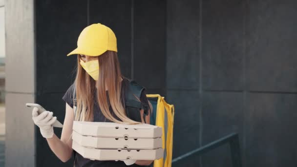 Mujer joven trabajadora de reparto en máscara médica de pie en la calle y sosteniendo cajas de cartón y el uso de teléfonos inteligentes. Mujer entregando comida y pizza usando gadgets para rastrear el orden en la calle de la ciudad. — Vídeos de Stock