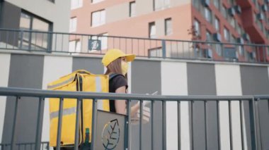 Beyaz, genç, güzel bir kadın, teslimatçı, kep ve tıbbi maske sokakta yürüyor ve içinde taze yiyecek olan bir paket taşıyor. Telefonlu güzel bir kurye. İnternet üzerinden mal teslimatı