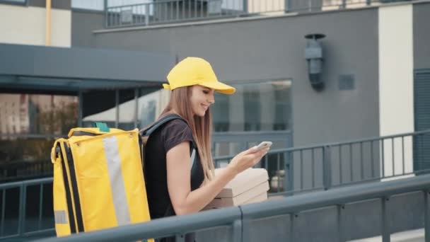 黒シャツの若い女性が街の通りで注文を追跡するためにガジェットを使って食べ物やピザを提供しています。宅配便オンラインアプリを使用してお支払いと配送先住所の追跡. — ストック動画