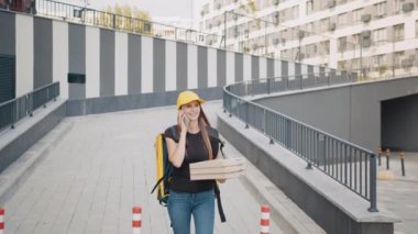 Beyaz kadın kurye bir müşteriyi telefonla arıyor. Çantalı bir kurye dışarıda müşterilere kutu getirir. Kurye telefonda konuşur ve caddede yürür.