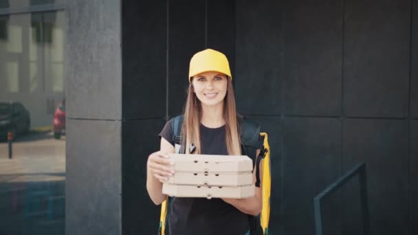 Női kézbesítő pózol pizzás dobozokkal. Pizzafutár szolgálat. A futárnő megmutatja a pizzát. Egy fiatal nő forró pizzát tart a dobozban. Élelmiszer-szállítási szolgáltatás női dolgozója a ház közelében. — Stock videók