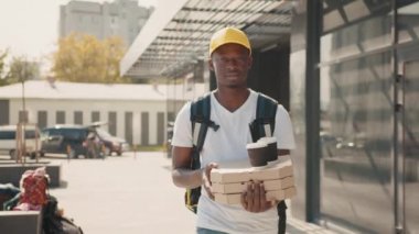 Modern evin dışında duran müşteriye kahve fincanı ve pizza getiren siyah, erkek kurye, siyah adam mutlu görünüyor, gülümsüyor, sarı üniforma giyiyor, şapka takıyor.