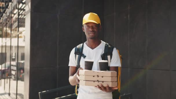 Joven repartidor africano en uniforme blanco con cajas de pizza, lonchera y café, mirando a cámara y sonrisa, listo para servir a los clientes clientes. Servicio de entrega, pedidos para llevar — Vídeo de stock